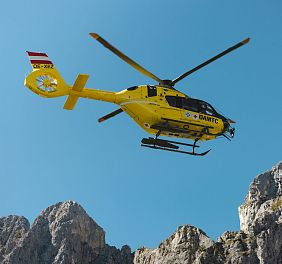 A yellow rescue helicopter soars above the rocky peaks of the Wilder Kaiser against a backdrop of clear blue sky, showcasing dynamic alpine rescue in action.