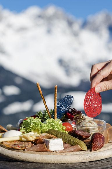 Ein Holzteller mit verschiedenen Käse- und Wurstsorten vor einer schneebedeckten Bergkulisse. Eine Hand greift nach einer Scheibe Wurst.
