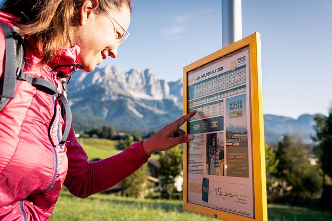 Eine Frau in outdoor Kleidung betrachtet ein Schild vor einer Berglandschaft mit grünen Wiesen und hohen Gipfeln im Hintergrund.