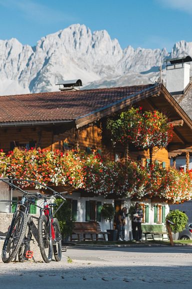 Zwei Radfahrerinnen sitzen mit Helmen auf einer Bank vor einer traditionellen Holzhütte mit Blumenschmuck. Im Hintergrund das imposante Bergmassiv Wilder Kaiser.