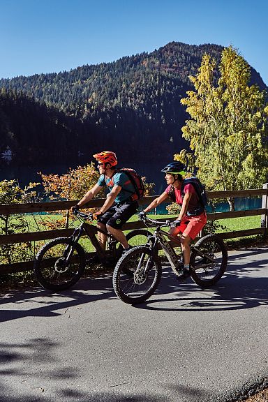 Zwei Fahrradfahrer auf einem Weg am See, umgeben von herbstlichen Bäumen und einem bewaldeten Hügel im Hintergrund, genießen die sonnige Landschaft.