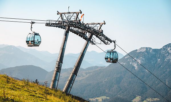 Moderne Gondelbahn fährt über grasbewachsene Hügel, mit beeindruckendem Bergpanorama im Hintergrund unter klarem, blauem Himmel.