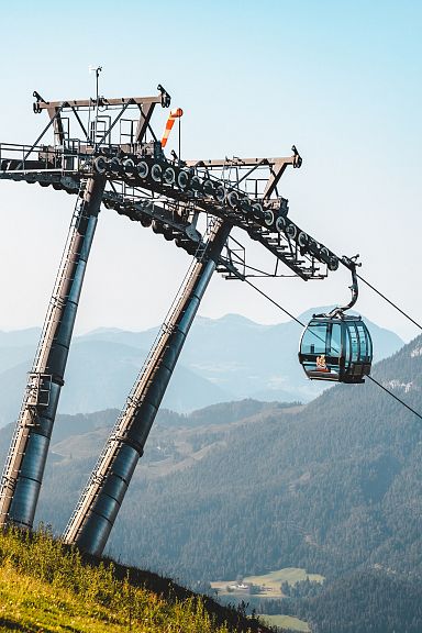 Moderne Gondelbahn fährt über grasbewachsene Hügel, mit beeindruckendem Bergpanorama im Hintergrund unter klarem, blauem Himmel.