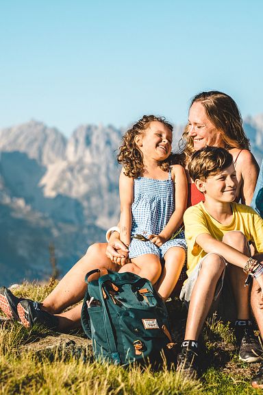 Eine vierköpfige Familie sitzt lachend auf einer Wiese vor einem imposanten Bergpanorama. Der Himmel ist klar und die Sonne scheint.