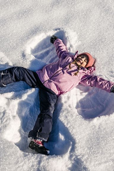 Ein Kind in rosa Jacke und schwarzer Hose liegt im Schnee und macht einen Schneeengel. Der Himmel ist nicht sichtbar, nur der weiße, glitzernde Schnee.