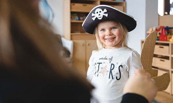 Kleines Mädchen mit Piratenhut lächelt in einem Spielzimmer mit bunten Boxen und Spielzeug. Es trägt ein T-Shirt mit der Aufschrift "Made of Stars".