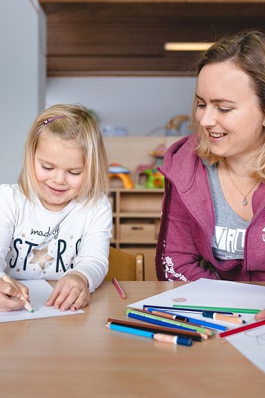 Eine Frau und ein kleines Mädchen zeichnen zusammen an einem Tisch. Sie sind entspannt und lächeln, umgeben von bunten Buntstiften und Zeichnungen.