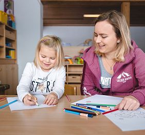 Eine Frau und ein kleines Mädchen zeichnen zusammen an einem Tisch. Sie sind entspannt und lächeln, umgeben von bunten Buntstiften und Zeichnungen.