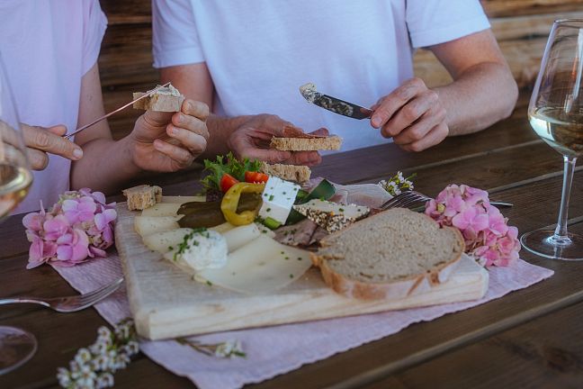 Zwei Personen genießen eine Jause mit Käse, Brot und Wein. Eine Holzplatte mit verschiedenen Käsesorten und Gemüse liegt auf einem Holztisch, umgeben von Blumen.