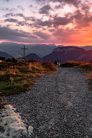 Sonnenuntergang in den Bergen mit einem Bergkreuz und einem schmalen Wanderweg im Vordergrund. Dramatische Wolken und warme Farben am Himmel.