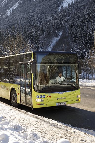 Yellow bus on a snowy road in the Wilder Kaiser region with snow-covered trees and mountains in the background.