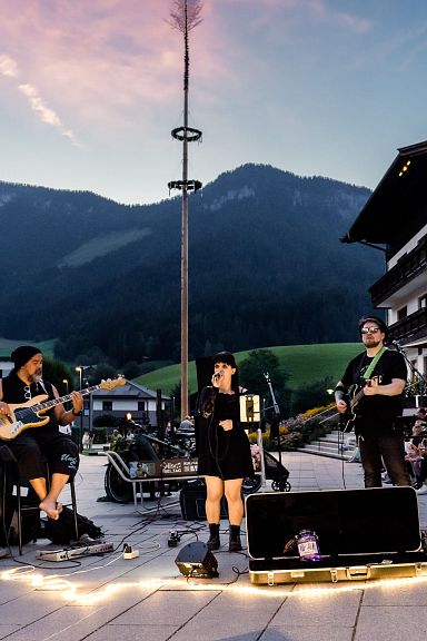 Eine dreiköpfige Band spielt bei Abenddämmerung im Freien, umgeben von traditionellen Gebäuden und Bergkulisse. Zuschauer sitzen entspannt vor der Bühne.