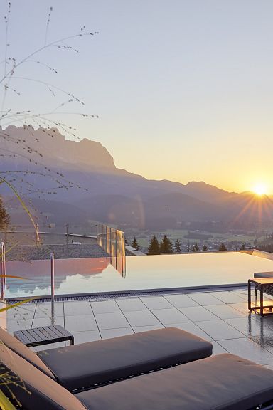 Terrace view with lounge chairs and a pool overlooking the Wilder Kaiser mountains at sunset, capturing a peaceful and scenic atmosphere.