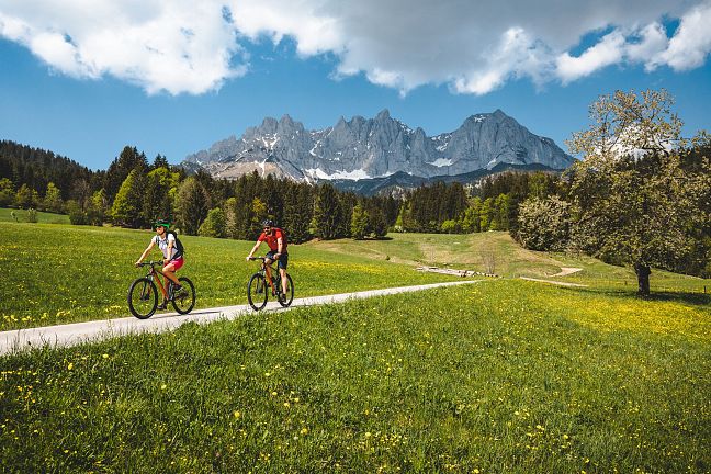 Zwei Personen fahren mit Fahrrädern auf einem Weg durch eine grüne Wiese, im Hintergrund ist ein beeindruckendes Bergmassiv mit blauem Himmel zu sehen.