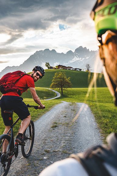 Two mountain bikers on a scenic trail, one in focus wearing a helmet, with the Wilder Kaiser mountains majestically rising in the background under a cloudy sky.
