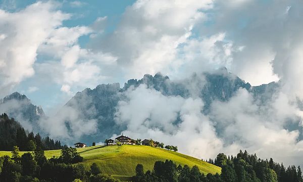View of the Wilder Kaiser mountains partially covered in clouds with green hills and a farmhouse in the foreground in Tirol.