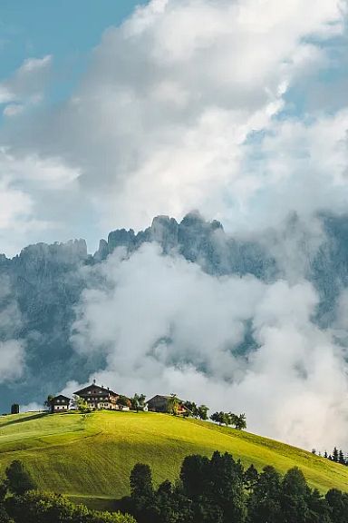 View of the Wilder Kaiser mountains partially covered in clouds with green hills and a farmhouse in the foreground in Tirol.