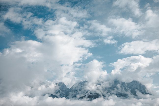 Blick auf eine Bergkette, teils von Wolken verhüllt, unter blauem Himmel. Die Spitzen der Berge ragen durch die Wolken hindurch, umgeben von dramatischem Licht.