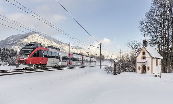 A train travels through a snowy landscape near a small chapel, set against the backdrop of the Wilder Kaiser mountains in winter.
