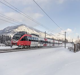 Ein roter Zug fährt im Winter durch verschneite Landschaften, vorbei an einer kleinen Kapelle, mit Bergen im Hintergrund unter einem klaren, blauen Himmel.