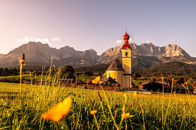 Idyllische Landschaft in Tirol mit einer Kirche im Vordergrund und dem imposanten Wilder Kaiser Gebirge im Hintergrund bei Sonnenuntergang.