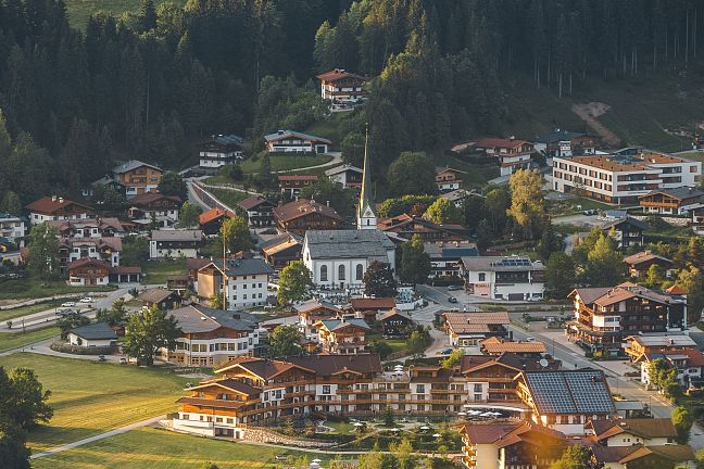 A scenic village in the Wilder Kaiser region, surrounded by green fields and mountains, under a clear sky, depicting rural life in Austria.