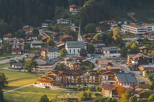 Luftaufnahme eines malerischen Dorfes in einer grünen Landschaft, umgeben von Wäldern und Hügeln, mit einer zentral gelegenen Kirche und verstreuten Häusern.