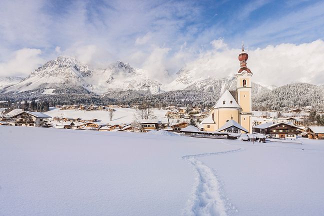 Winterlandschaft mit schneebedecktem Dorf und Kirche im Vordergrund, umgeben von majestätischen Bergen unter bewölktem Himmel.