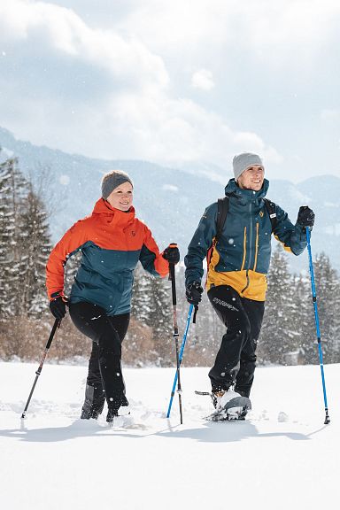 Zwei Personen in Winterkleidung machen eine Schneeschuhwanderung durch eine verschneite Landschaft mit Bäumen und Bergen im Hintergrund.