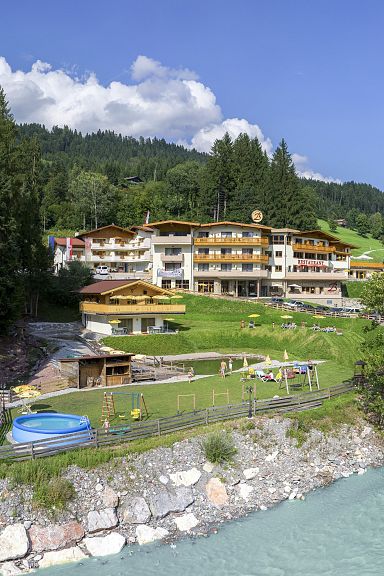 Ein Hotel liegt idyllisch in den Bergen mit einem Fluss im Vordergrund. Es ist umgeben von üppigem Grün und Bäumen unter einem blauen Himmel mit Wolken.