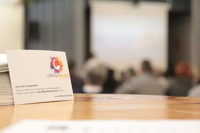 A focused business card with blurred figures in the background at a conference or meeting setting.