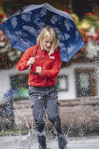 Ein Kind in roter Jacke springt fröhlich durch eine Pfütze mit Regenschirm, während im Hintergrund bunte Blumen und ein traditionelles Holzhaus zu sehen sind.
