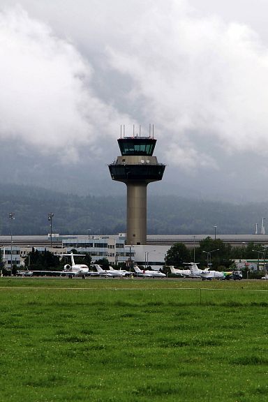 Ein Flughafen mit einem hohen Kontrollturm und geparkten Flugzeugen. Im Hintergrund sind bewaldete Berge und Wolken am Himmel zu sehen.
