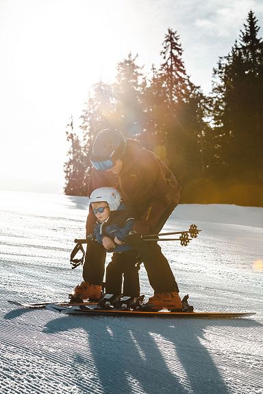A child and adult skiing on a sunlit slope in Wilder Kaiser region, surrounded by trees, capturing a serene winter landscape.