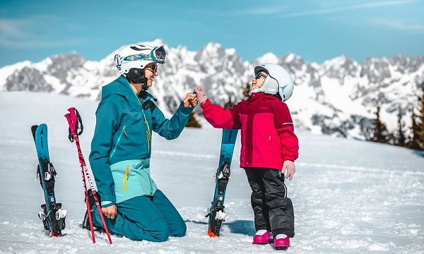 Erwachsener und Kind im Schnee mit Skiausrüstung vor beeindruckendem Alpenpanorama, lächeln sich an. Die Szene wirkt winterlich und fröhlich.