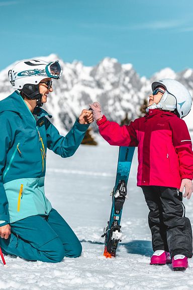 Erwachsener und Kind im Schnee mit Skiausrüstung vor beeindruckendem Alpenpanorama, lächeln sich an. Die Szene wirkt winterlich und fröhlich.