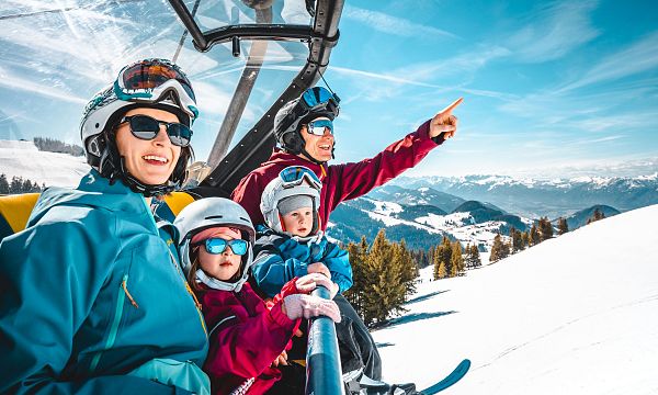 Familie in Skikleidung sitzt auf einem Sessellift in schneebedeckter Berglandschaft. Kinder tragen Helme, während der Erwachsene in die Ferne zeigt.