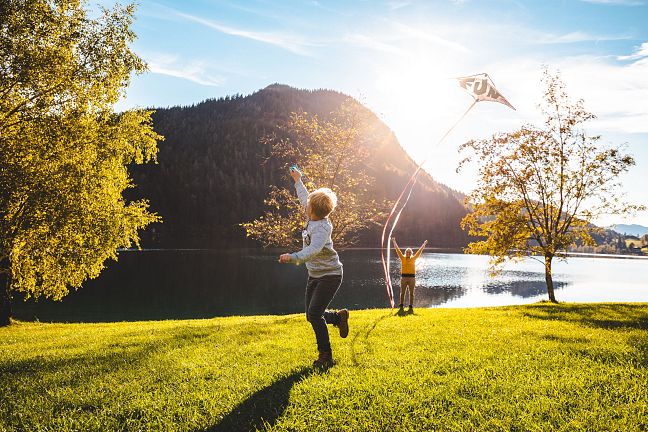 Zwei Kinder spielen auf einer Wiese am See, während die Sonne über die Berge scheint. Eines der Kinder lässt einen Drachen steigen. Eine malerische Herbstlandschaft.