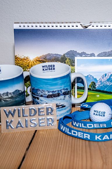 Ein Set Wilder-Kaiser-Souvenirs: Zwei Tassen mit Bergmotiven, ein Kalender, Postkarten, ein Schlüsselband, ein Stift und eine Verpackung auf einem Holztisch.