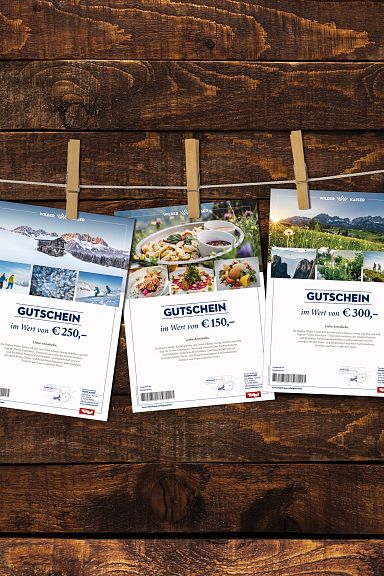 Drei Gutscheine hängen mit Klammern an einer Leine vor einer rustikalen Holzwand. Jeder Gutschein zeigt verschiedene Fotos und Werte in Euro.