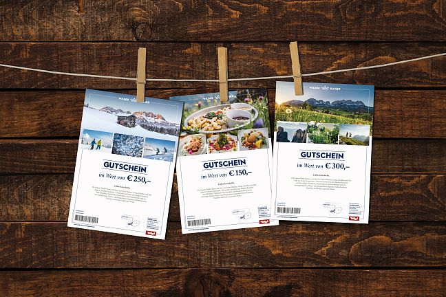 Drei Gutscheine hängen mit Klammern an einer Leine vor einer rustikalen Holzwand. Jeder Gutschein zeigt verschiedene Fotos und Werte in Euro.