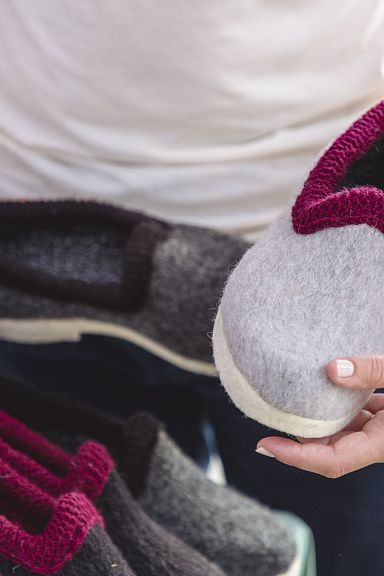 Personen halten warme, handgefertigte Filzhausschuhe in Grau und Rot. Ideal für kalte Tage. Detailaufnahme von Händen und Schuhen auf einem Markt.