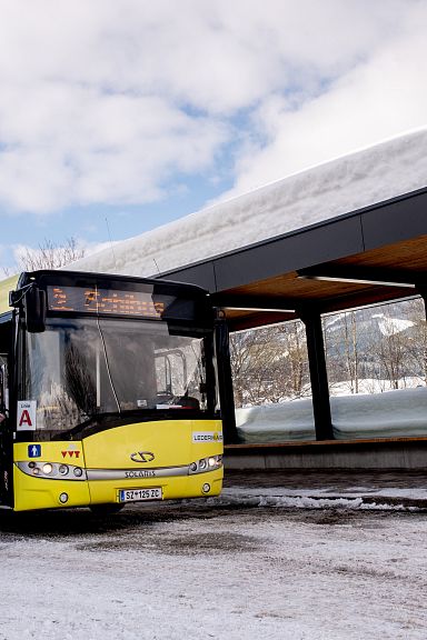 Ein gelber Bus hält an einer verschneiten Haltestelle. Eine Person steigt mit Skiern ein. Im Hintergrund verschneite Berge unter einem bewölkten Himmel.