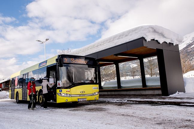 Ein gelber Bus hält an einer verschneiten Haltestelle. Eine Person steigt mit Skiern ein. Im Hintergrund verschneite Berge unter einem bewölkten Himmel.