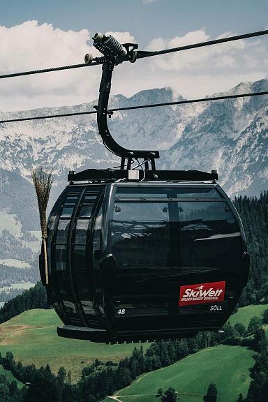 Cable cars traverse a verdant landscape, with towering peaks of the Wilder Kaiser in the distance, under a partly cloudy sky.