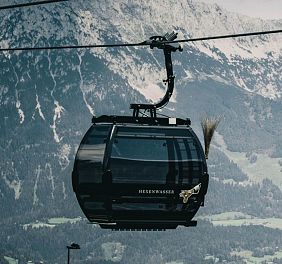 Zwei schwarze Gondeln der SkiWelt-Seilbahn vor alpiner Kulisse, mit schneebedeckten Bergen und grünen Hügeln im Hintergrund, in der Region Wilder Kaiser.
