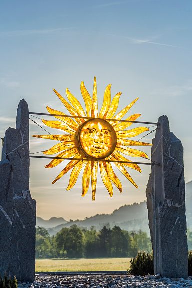 Kunstinstallation: Eine eindrucksvolle Skulptur aus großen Steinen, die einen stilisierten, goldenen Sonnenaufgang über einer idyllischen Berglandschaft darstellt.