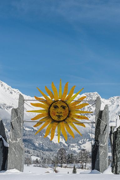 Sun-themed art installation with snowy peaks of Wilder Kaiser in the background, contrasting against a clear blue sky and white snow, depicting winter beauty.