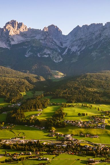 Panoramablick auf das imposante Bergmassiv des Wilden Kaisers in Tirol, eingerahmt von grünen Wiesen und malerischen Dörfern unter einem klaren blauen Himmel.