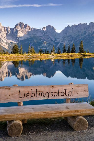 Holzbank mit Aufschrift 'Lieblingsplatzl' vor einem See, der die dramatischen Berge des Wilden Kaisers in Tirol bei klarem Himmel spiegelt.